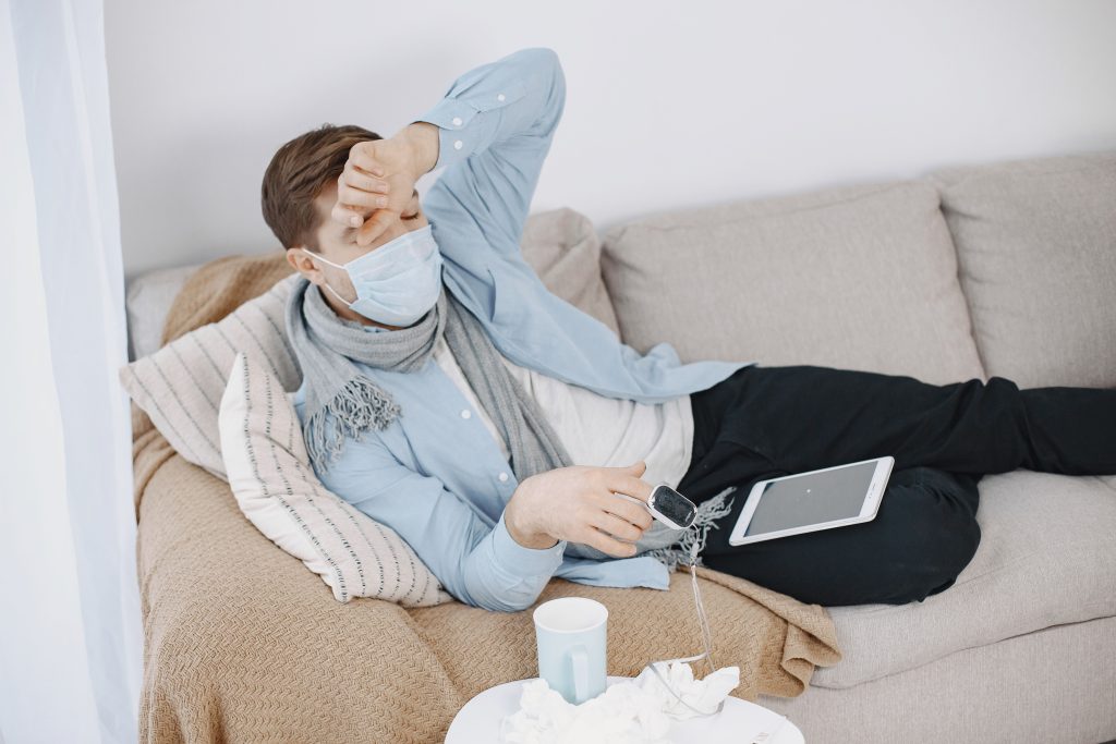 Man in mask resting on sofa with pulse oximeter and tablet, conveying illness at home.